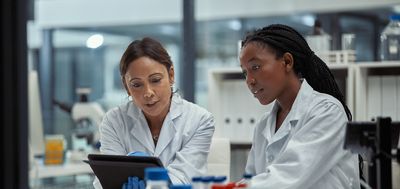 Two women in lab coats sit at a lab bench side-by-side viewing a tablet screen and conversing