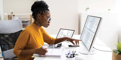 A young, Black female lab manager sits at her desk going through her lab's budget on a large computer monitor. She is wearing a yellow sweater and glasses.